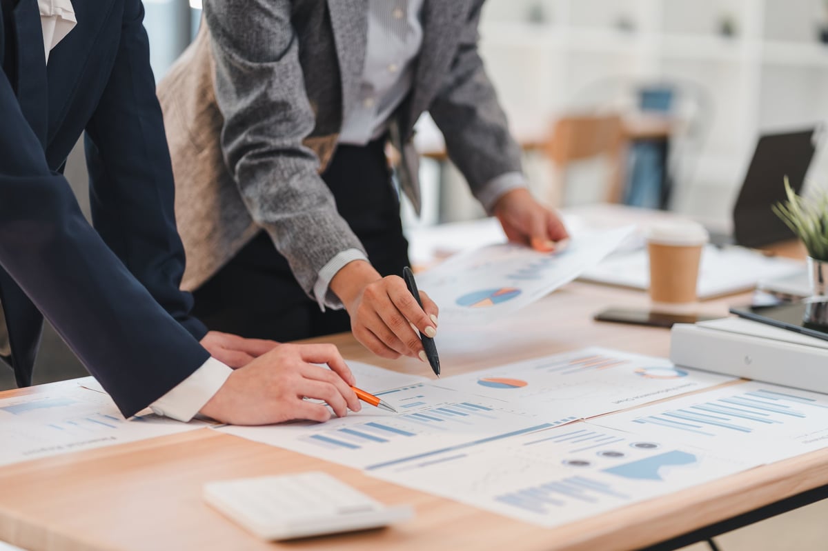 Two business professionals analyzing financial charts and graphs on wooden desk in modern office setting, collaborating on project